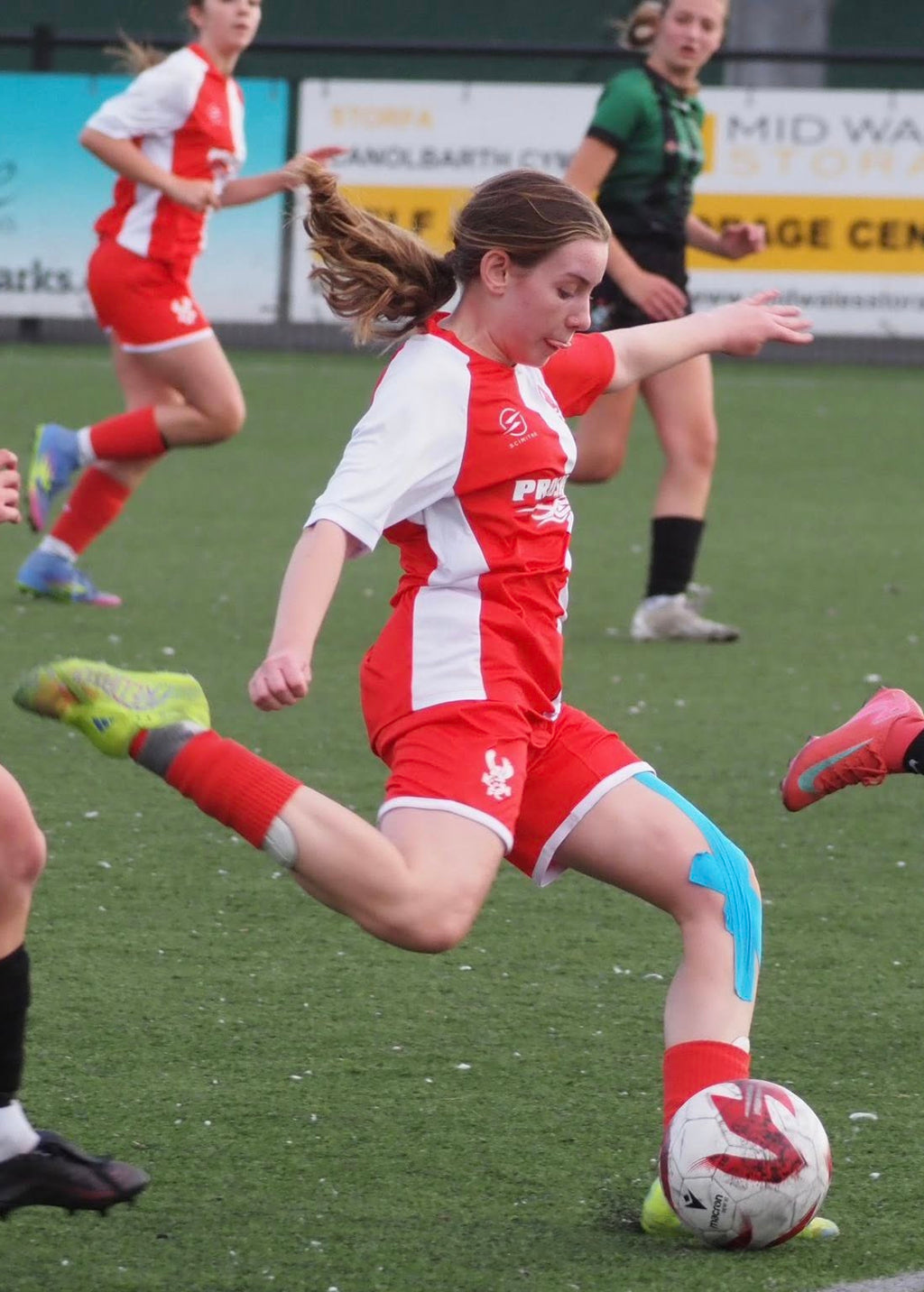 Young female soccer player in red and white uniform kicking a ball on a field with other players.