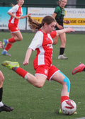 Young female soccer player in red and white uniform kicking a ball on a field with other players.