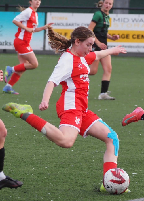 Young female soccer player in red and white uniform kicking a ball on a field with other players.