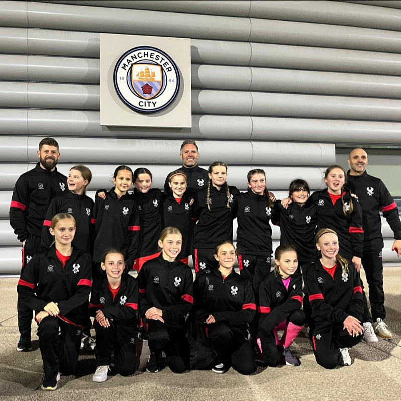 Group of people in black and red sports uniforms posing in front of a Manchester City logo.
